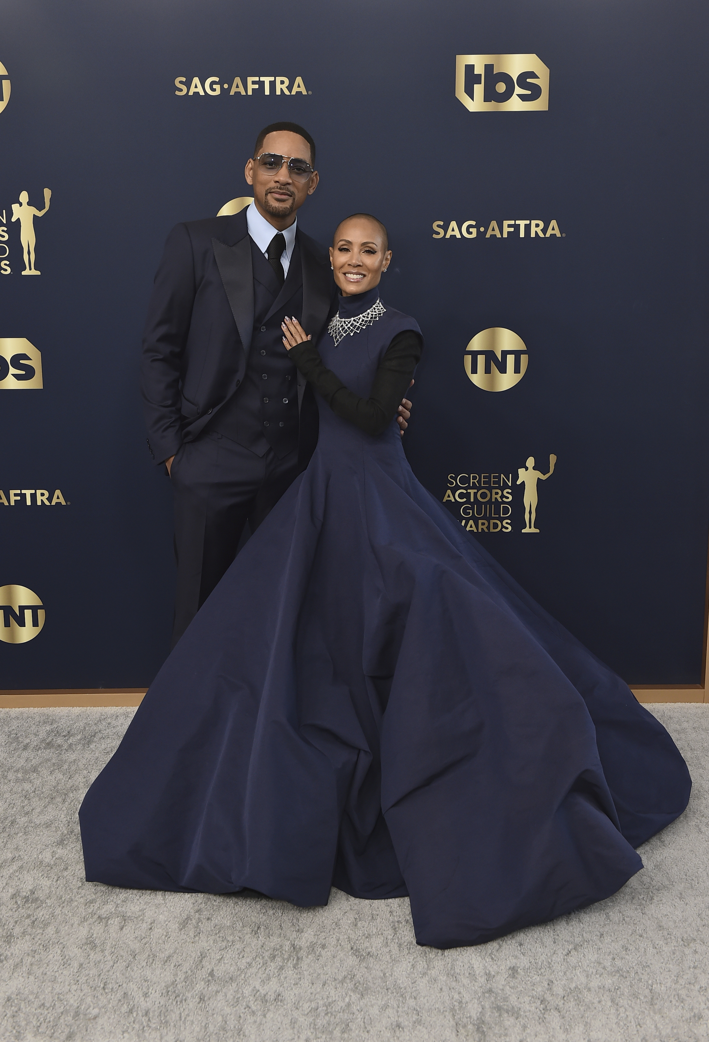 Will Smith wears a navy suit with sunglasses, Jada Pinkette Smith wears a navy floor length gown with long black sleeves