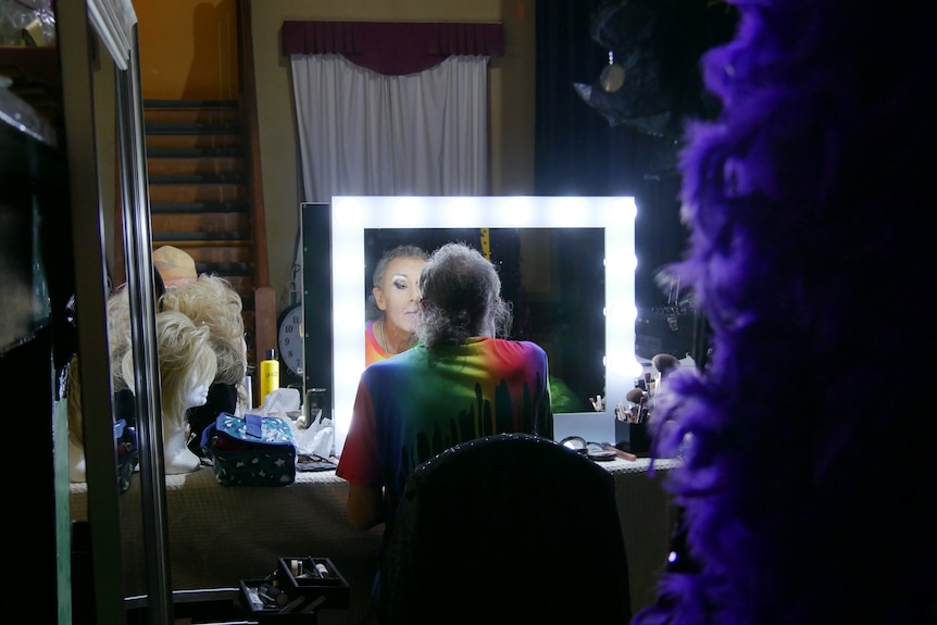 A man sits with his back to the camera in front of a mirror bordered with bright lightbulbs, with make-up and a wig. 