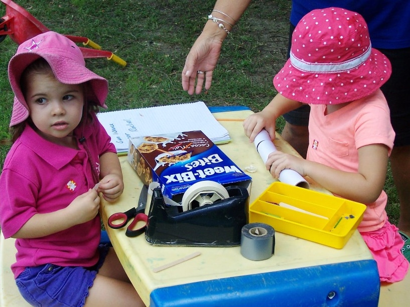 preschool aged girls sit at a table with cardboard, paper and recyclables making toy guns