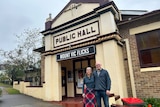 A man and a woman stand in front of an old public hall with a sign reading 'Mount Vic Flicks' 