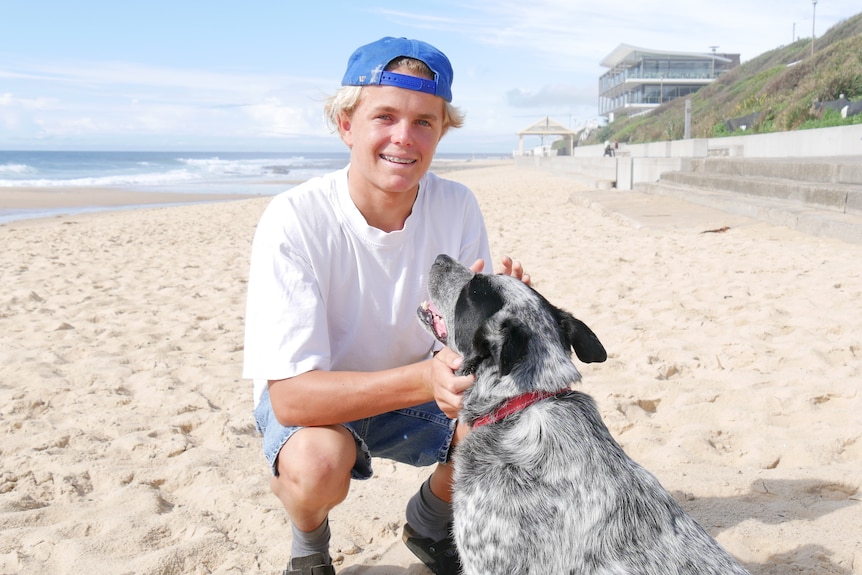 Manning squats down, patting his dog in front of him, smiling, with the beach behind him.