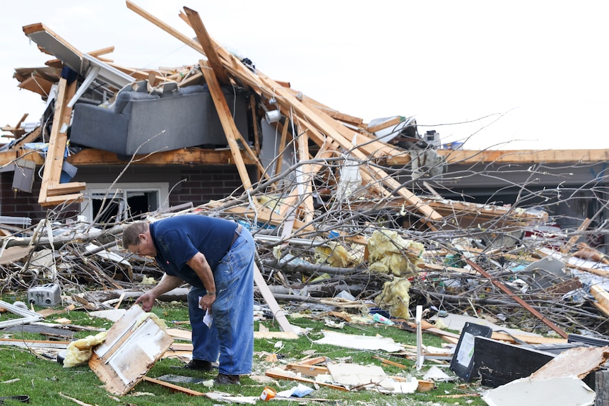 A man picking up a piece of debris in front of a destroyed house. 