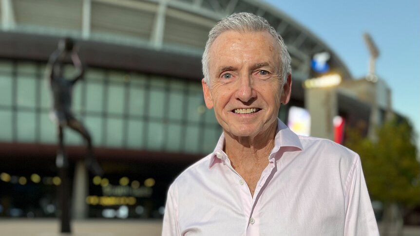 A man in a white shirt and with grey hair stands outside a football stadium.