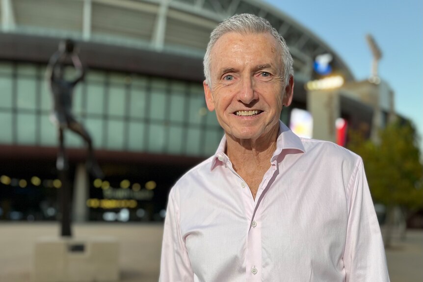 A man in a white shirt and with grey hair stands outside a football stadium.