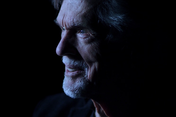 An older man with white hair and facial hair smiles and is lit with soft light in dark room.