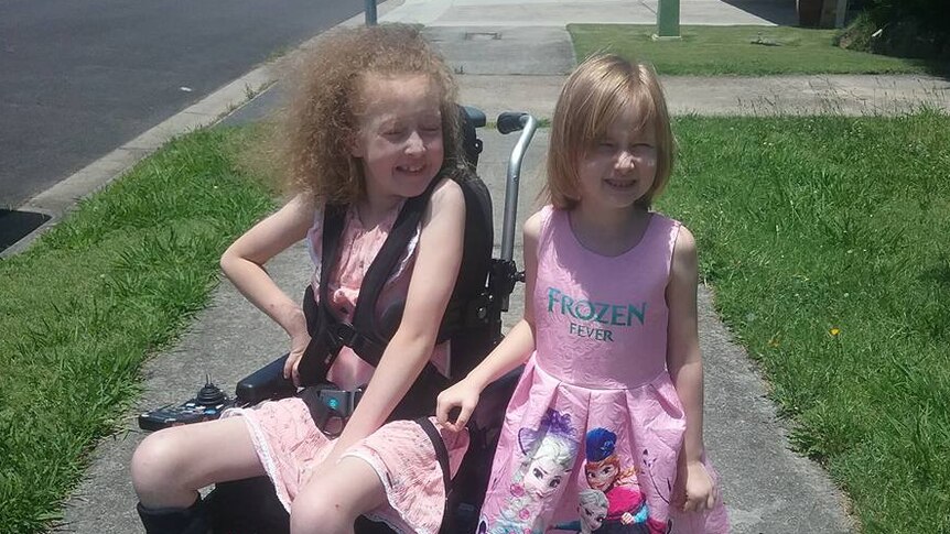 Two young girls, one in a wheelchair, smile together as they sit/stand on a footpath with green grass either side.