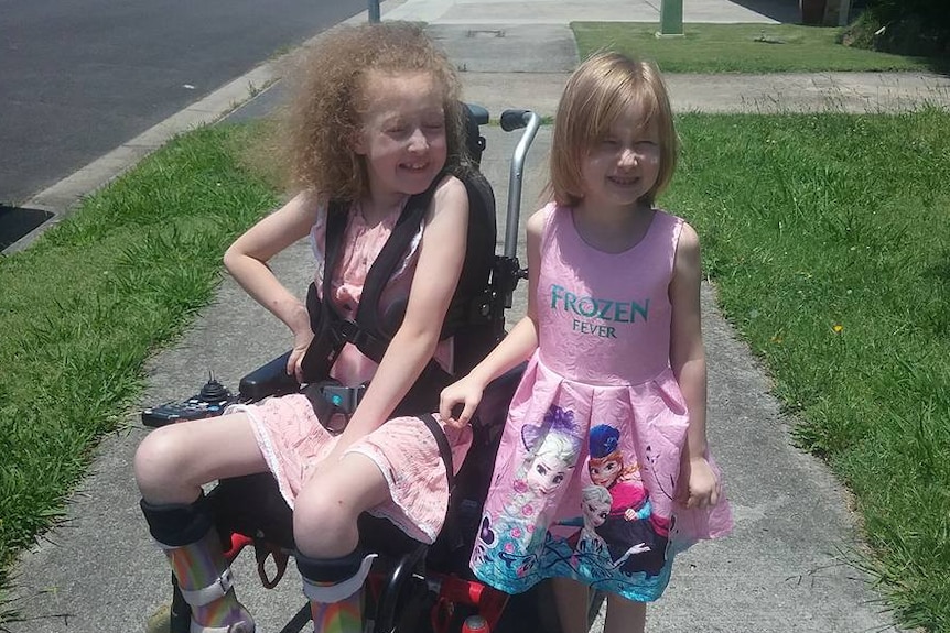 Two young girls, one in a wheelchair, smile together as they sit/stand on a footpath with green grass either side.