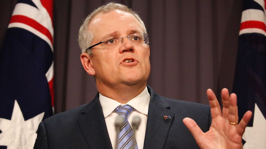 Liberal MP Scott Morrison speaks at a press conference with hand raised and two Australian flags in background.