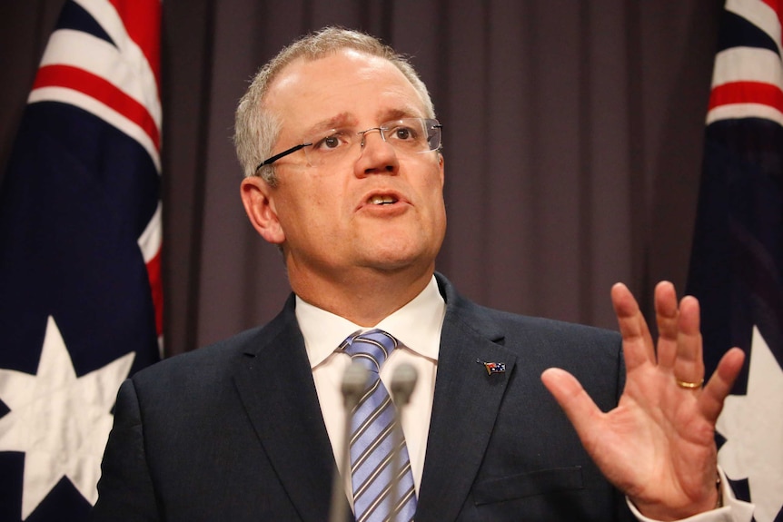 Liberal MP Scott Morrison speaks at a press conference with hand raised and two Australian flags in background.