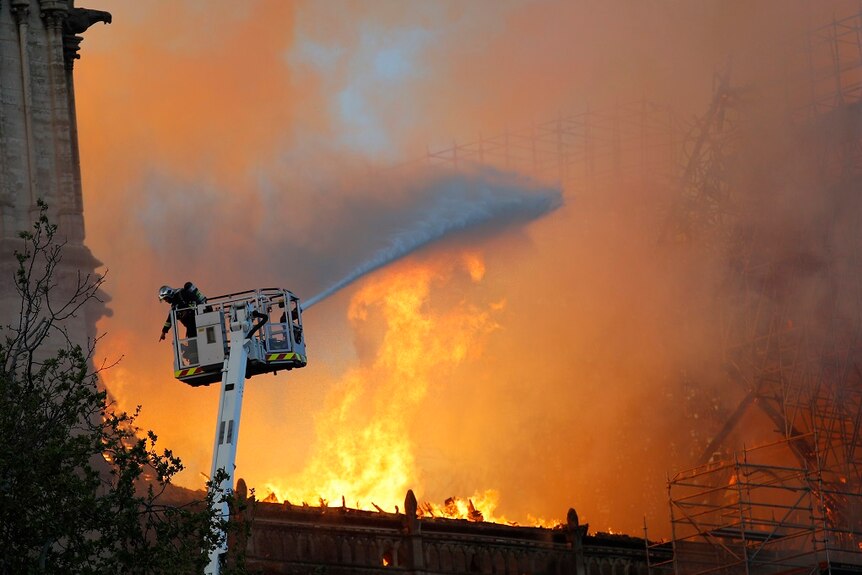 A firefighter gestures at a hose while in a cherry picker as large flames rise in the background