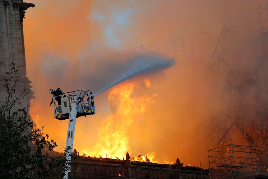 A firefighter gestures at a hose while in a cherry picker as large flames rise in the background