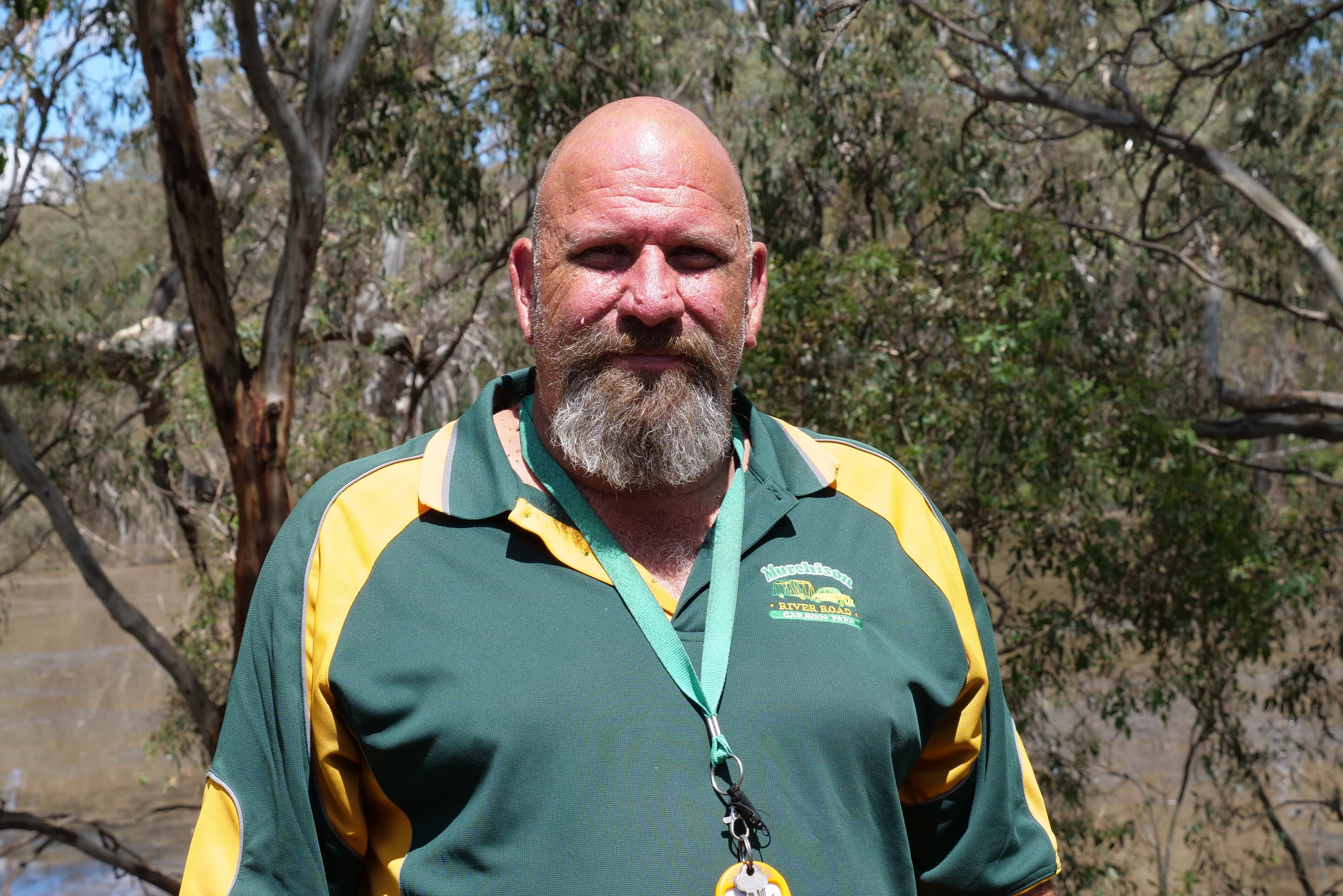 A man with bald head and beard wearing a green shirt standing in front of a tree.