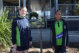 Two children stand either side of a small tree in front of a school demountable