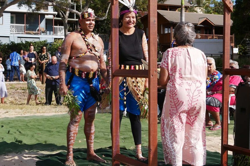 A man and woman wearing traditional Aboriginal outfits stand in front of an arch while a crowd behind them watches