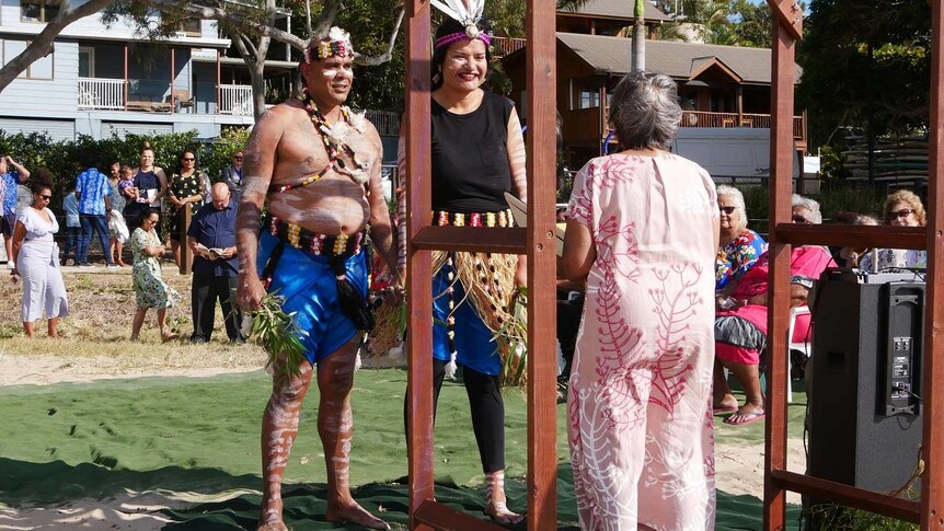 A man and woman wearing traditional Aboriginal outfits stand in front of an arch while a crowd behind them watches.i