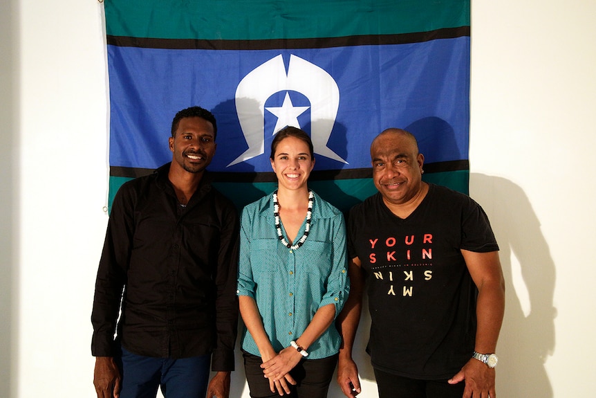 A Caucasian woman stands between two Indigenous Australians in front of a Torres Strait Island flag.