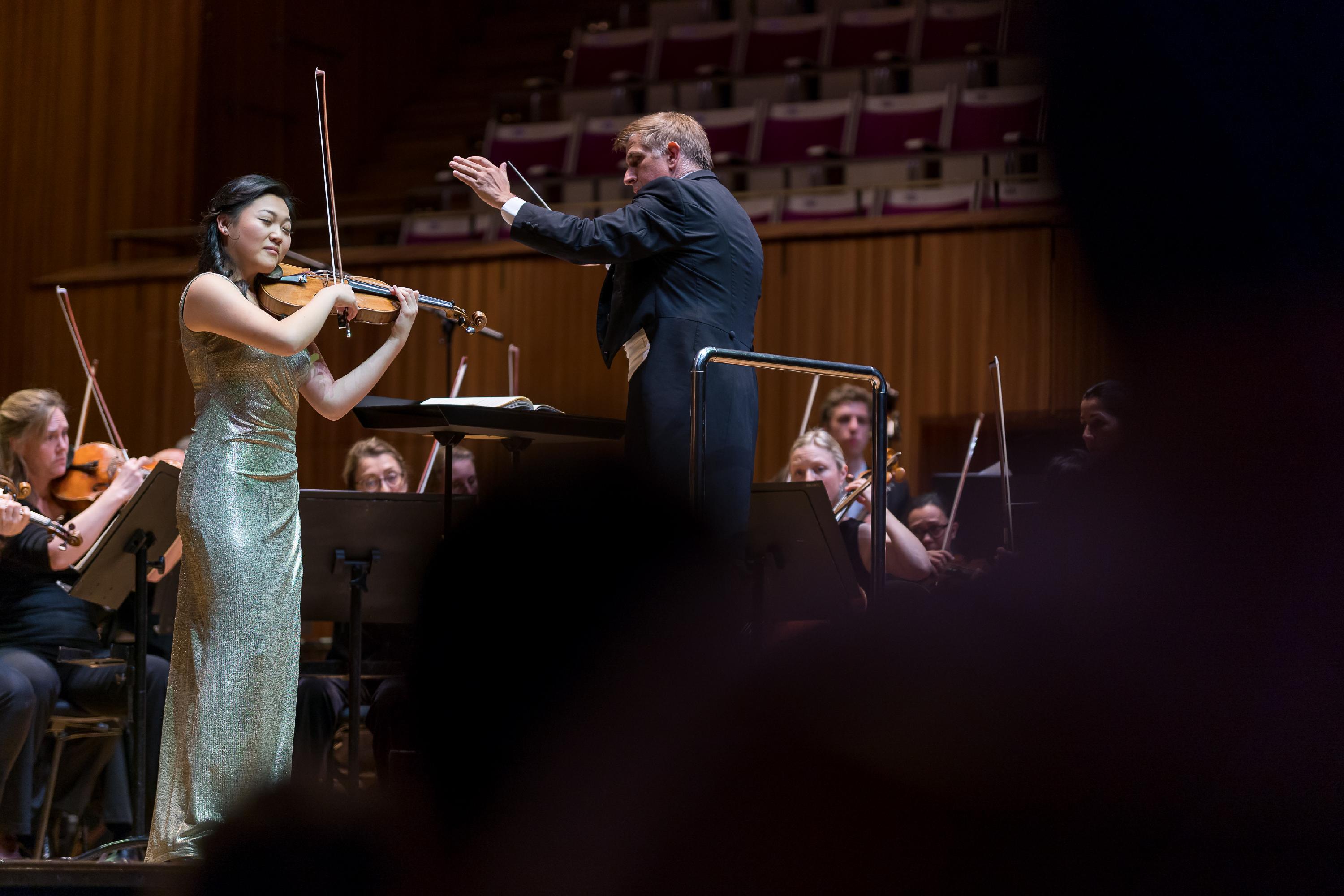 A violinist stands at the front of an orchestra in a concert hall, playing alongside the conductor who is waving his arms