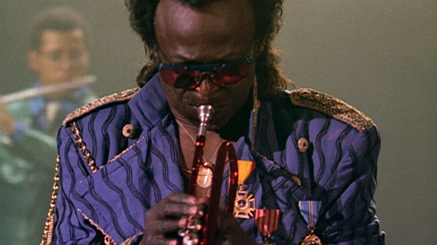 Miles Davis wears a purple jacket and looks down as he blows a trumpet