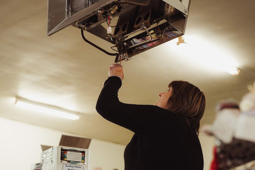 A woman withbrown hair dressed in a black shirt stands on a chair adjusting a ceiling exhaust inside.