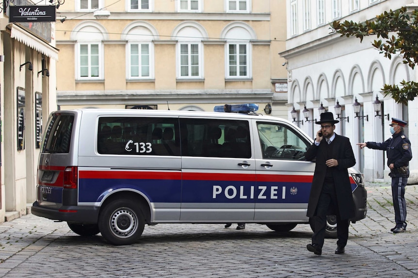 A rabbi in traditional black garb walks in front of a police van & officer in cobble-stoned square.