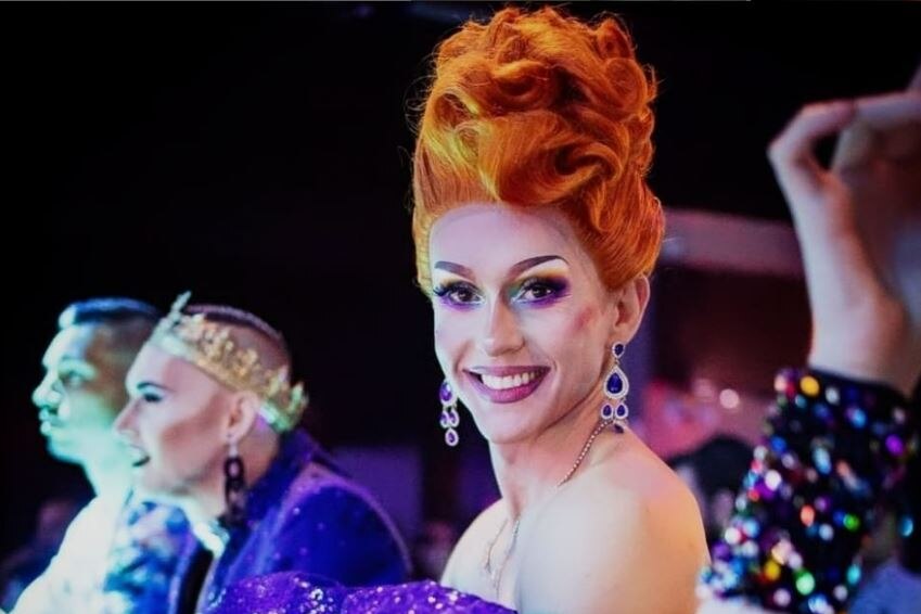 A drag performer with bright red hair smiles at the camera.