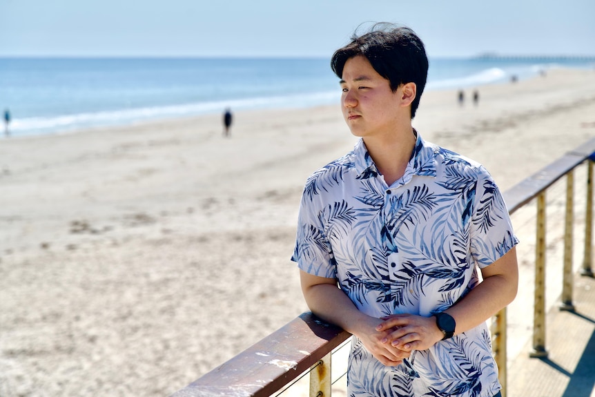 A man wearing a blue and white button up shirt leans his elbow on a fence and looks out over the beach