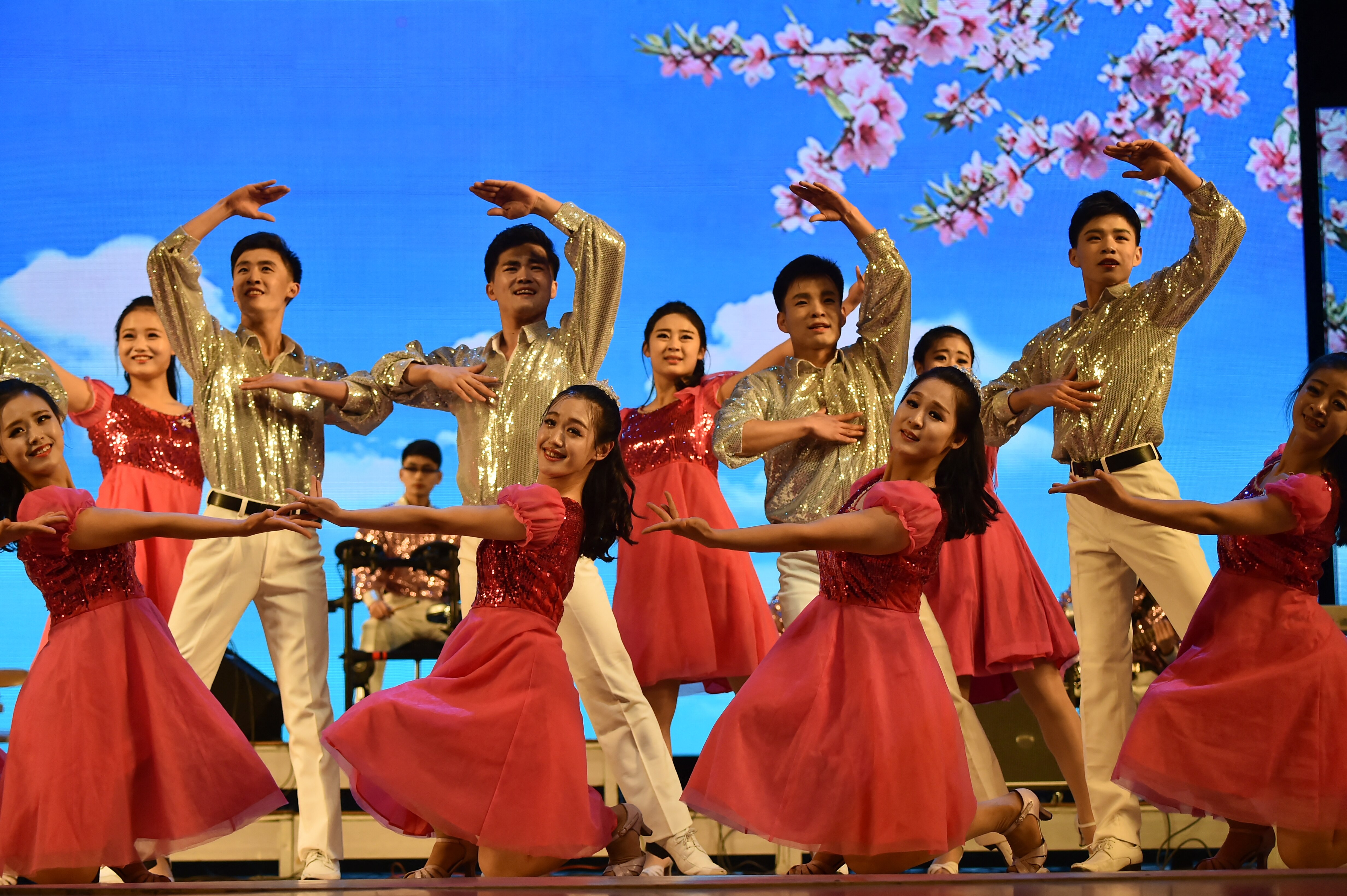 Women in red dresses and men in golf suits dance on stage with cherry blossoms in the background. 