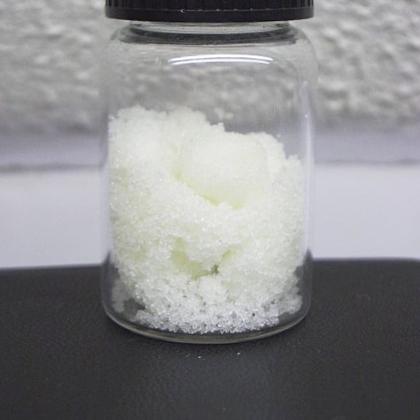 You view a small glass vial of white crystals sitting on a black benchtop against a white textured wall.