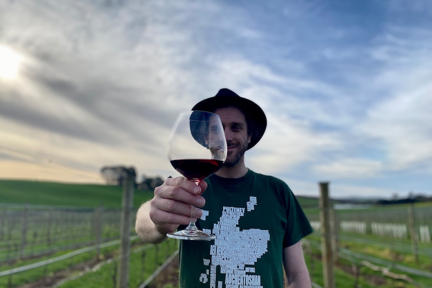 Man in black hat holds wine glass up to the camera, covering half his face