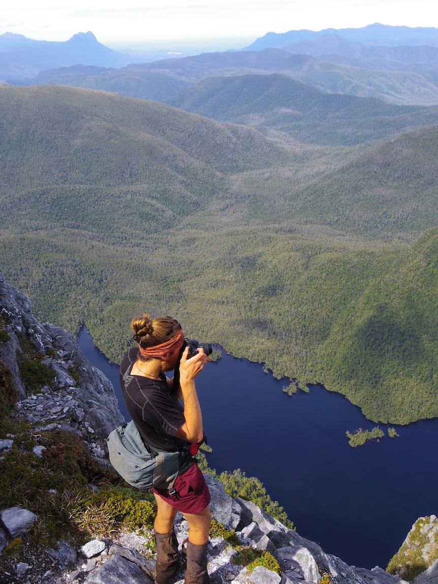 Picture of a man standing on a mountain overlooking a lake taking a photo