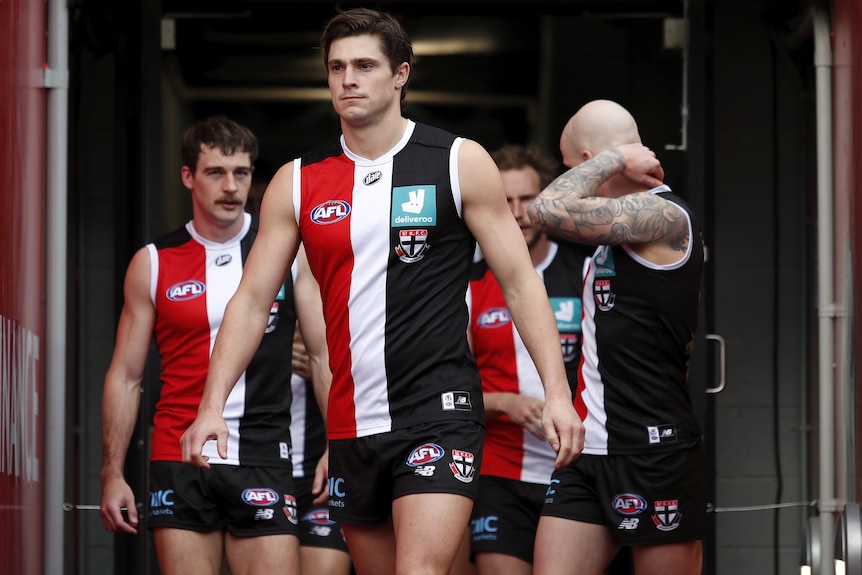 An AFL captain in red, black and white stands in the player's race before a game with his team behind him. 