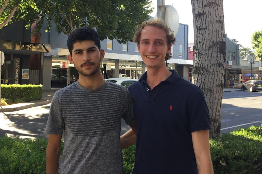 A man with black hair stands next to a man who has blonde hair and is smiling. They are on a street with trees and shops.