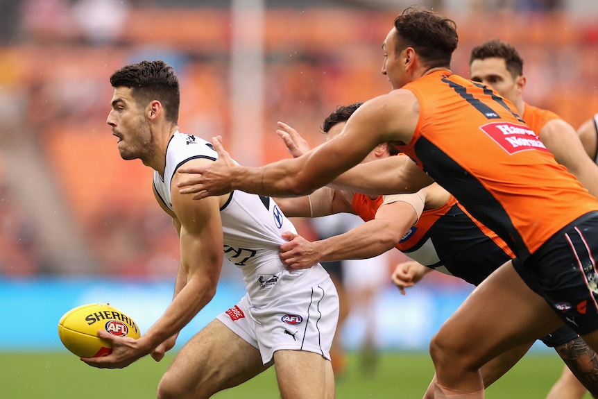 A Carlton player puts his fist to the ball to hand-ball as defenders grab him by the shirt and the shoulder.