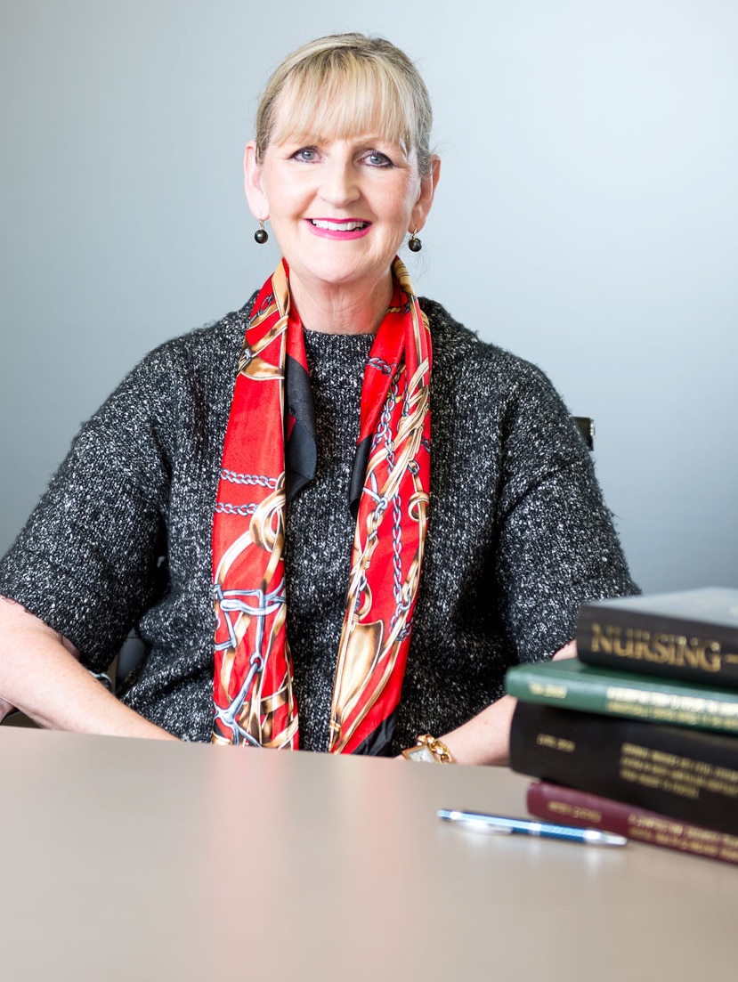 Lady in red scarf smiling sits at desk with stack of academic books on desk in foreground