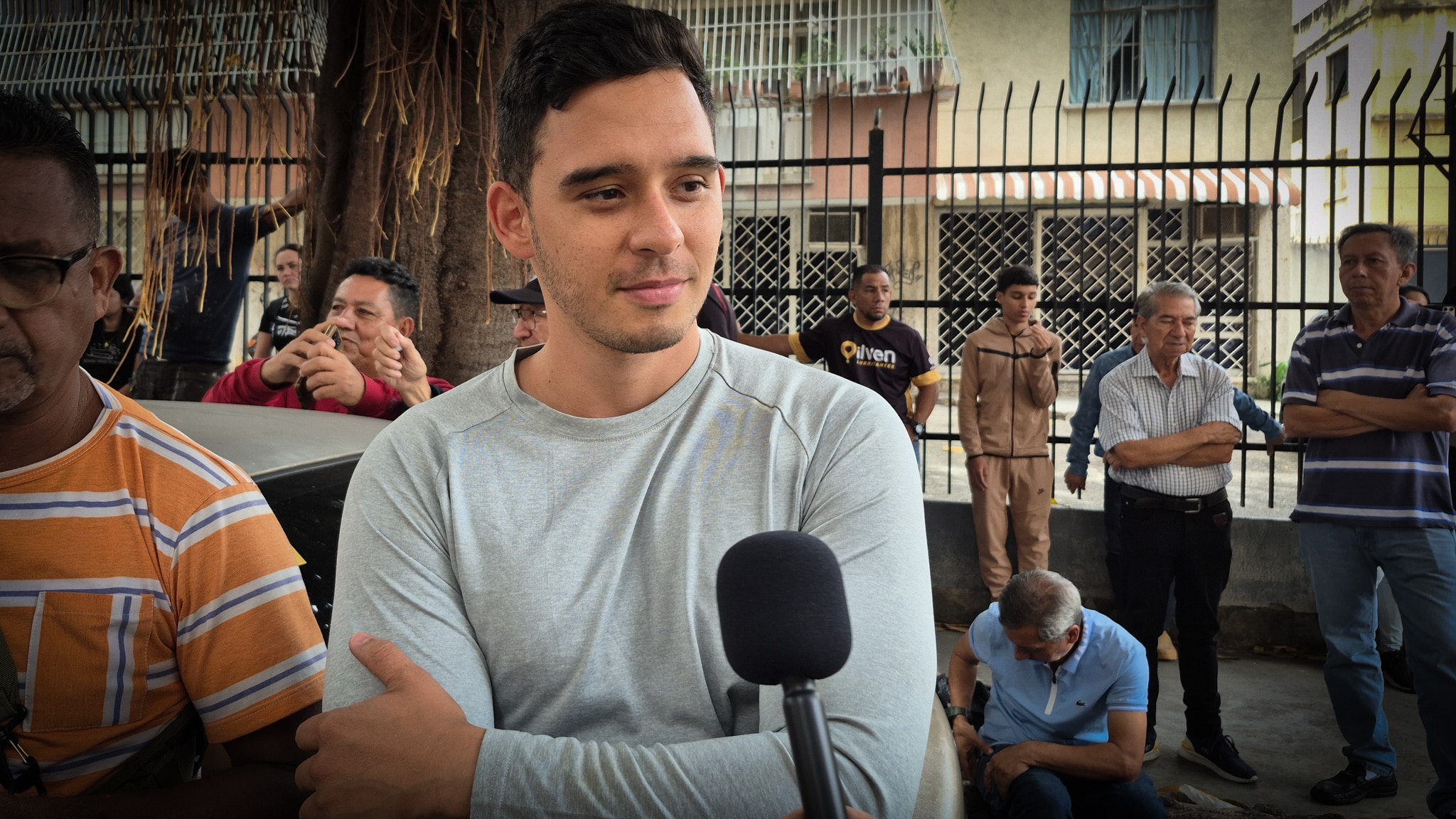 A close up of a man wearing a grey shirt speaking near other men on the street.