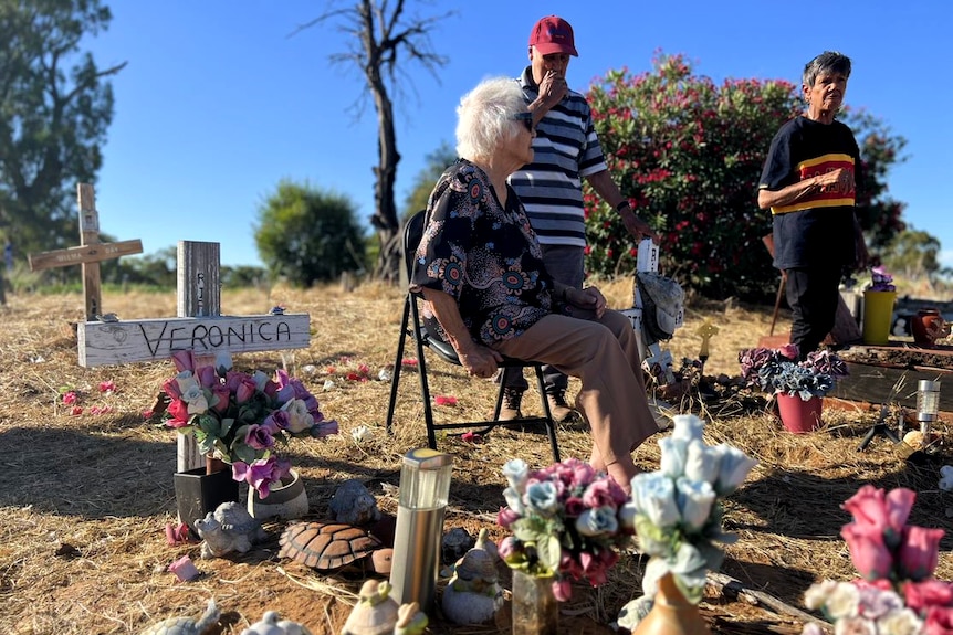 Three people gather around a white cross labelled "Veronica", with flowers and turtle statues over the site.