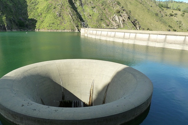 A giant concrete drain shown with its rim above the surface of a lake, with a dam wall and green hills behind it