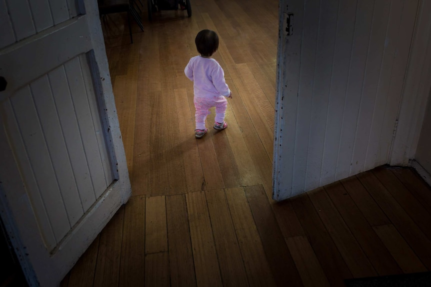 Tia, dressed in pink and wearing sandals, walks alone on floorboards.