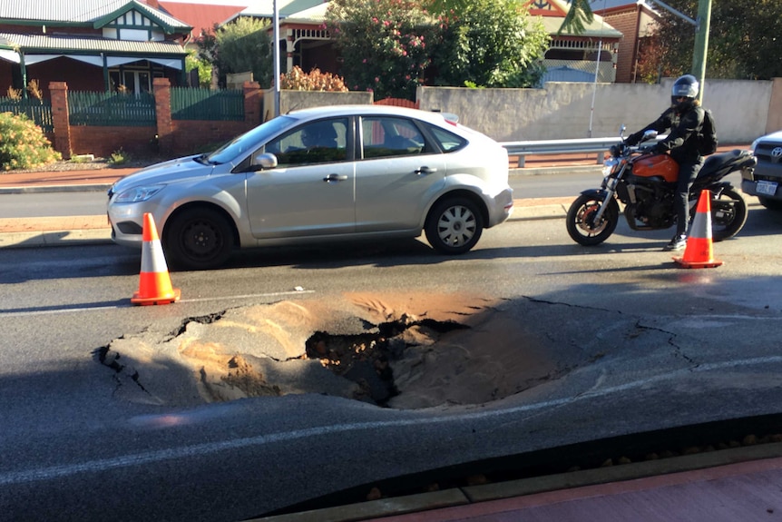 A sink hole in a road in t e foreground, with a car and motorbike.