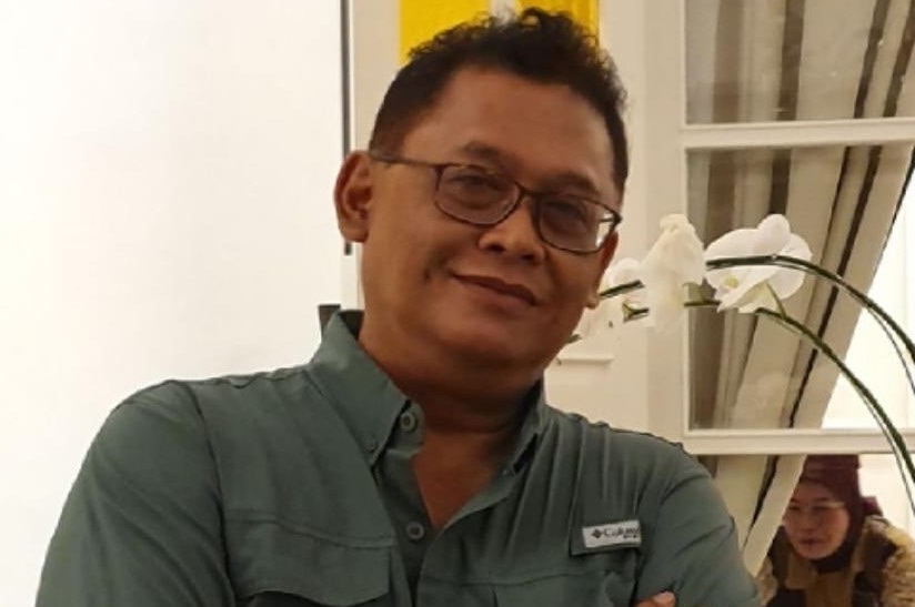 Pandu Riono poses with his arms crossed, standing right next to some white flowers.