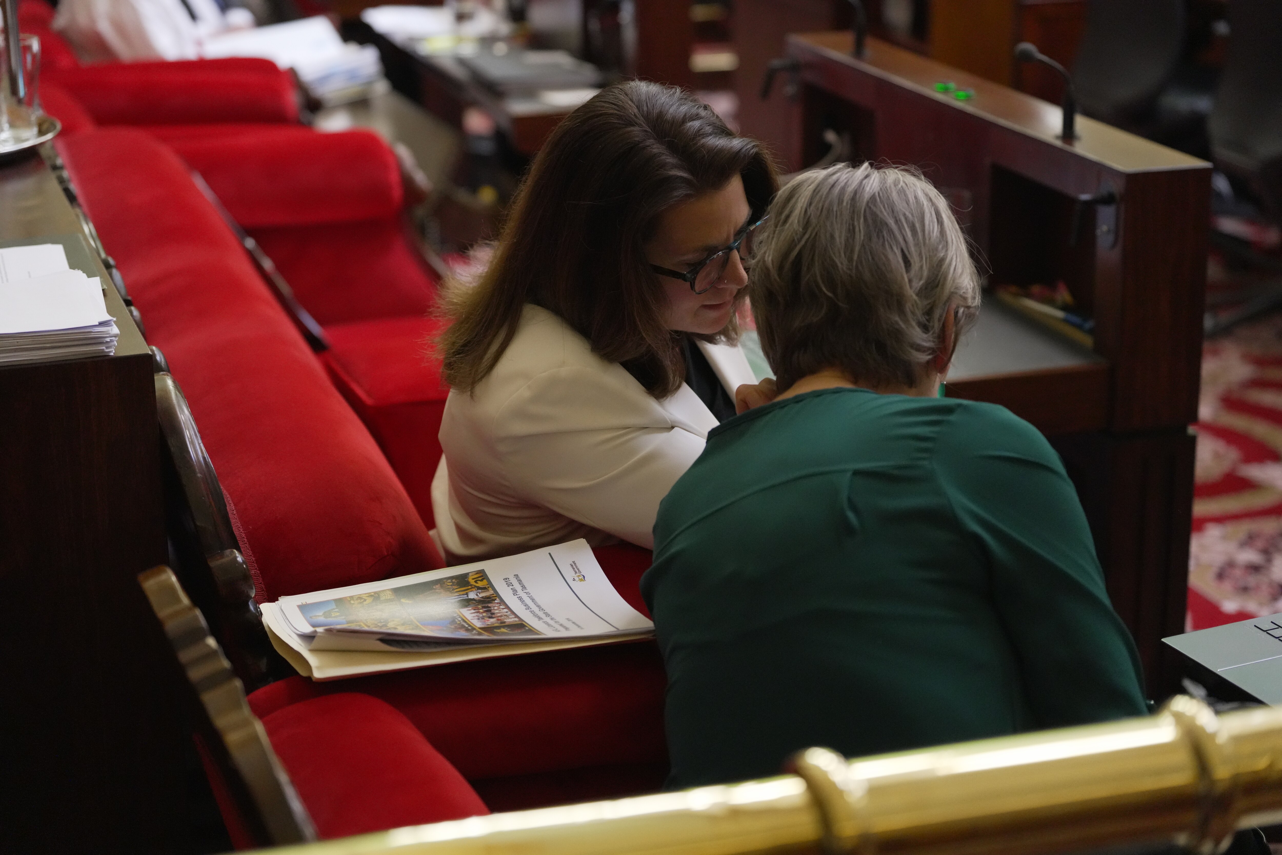 Two women sitting in red chairs lean in and talk to each other.