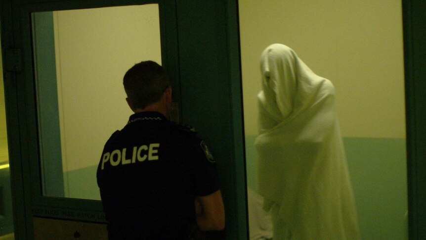 Prisoner stands inside a cell with a sheet over their head as an officer stands outside.