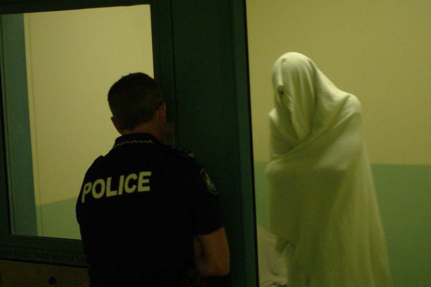 Prisoner stands inside a cell with a sheet over their head as an officer stands outside.