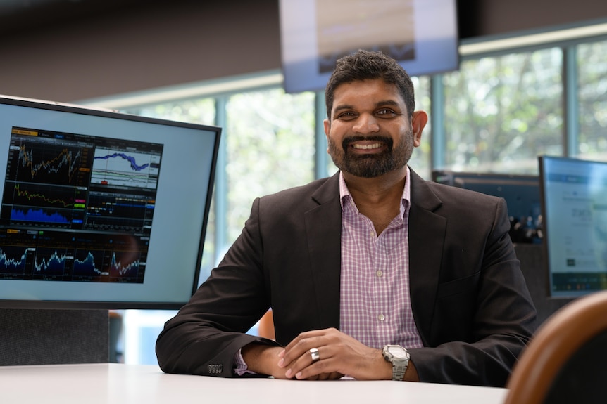 A smiling man with a dark, neat beard sits at a desk wearing a blazer.