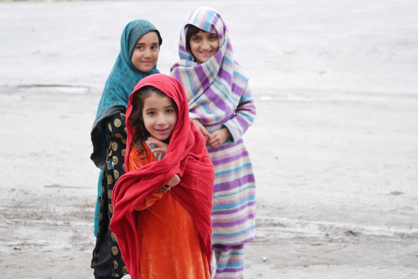 Three young Afghan girls smile for a photo while standing on a street.