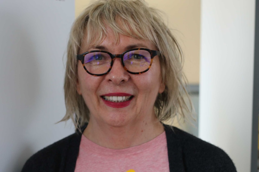 Midshot of a woman with blond hair and glasses, smiling, in front of a white wall.