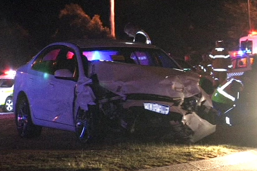 A shot of a white car with a smashed front end surrounded by police cars and ambulances.