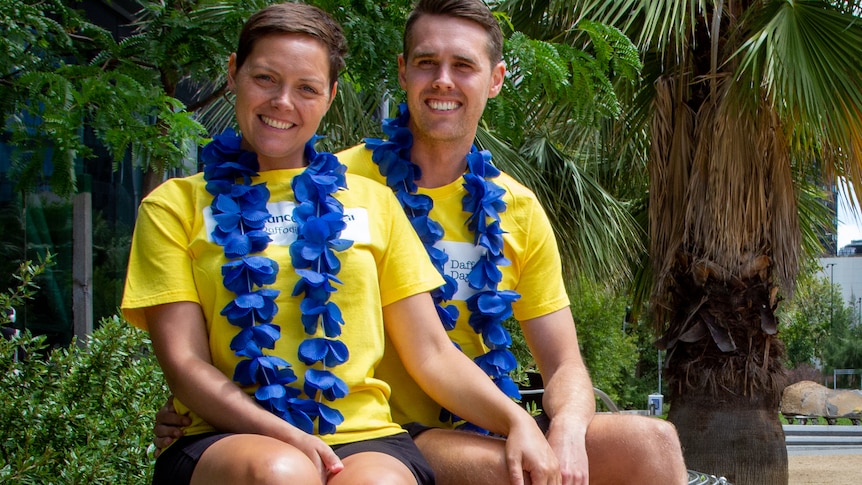 A young couple in matching cancer council t-shirts sitting outside on a metal chair.