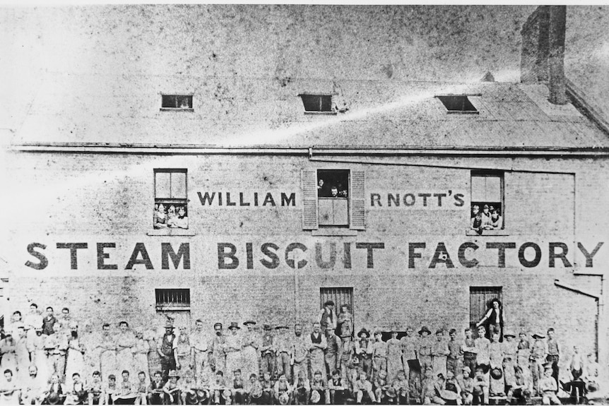 A black and white photo of a brick building with the words William Arnott's Steam Biscuit Factory on it, with workers in front.