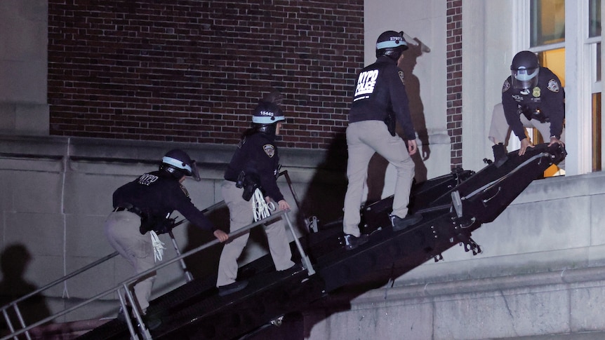 Three riot police climb up a ramp to a building, a fourth police officer holds it down